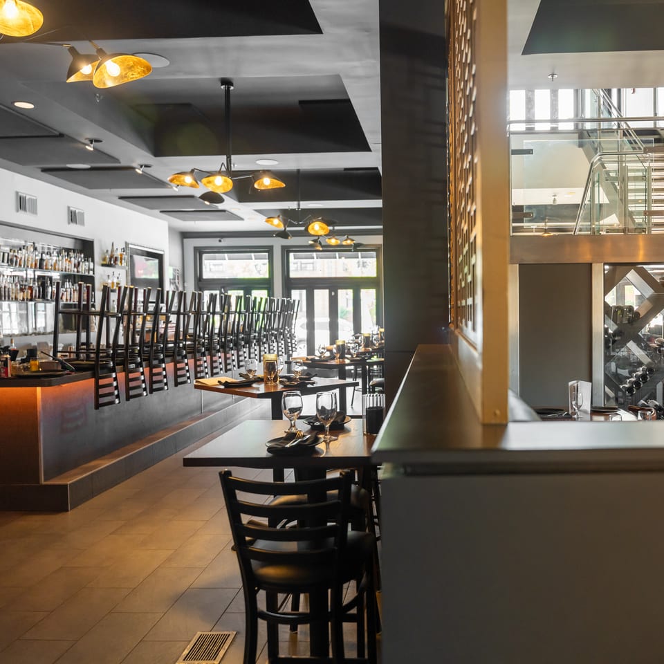 Modern restaurant interior featuring a bar with stacked chairs, set dining tables, and a glass staircase.