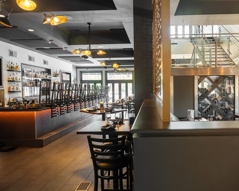 Modern restaurant interior with a bar, stacked chairs, set dining tables, and a glass staircase.