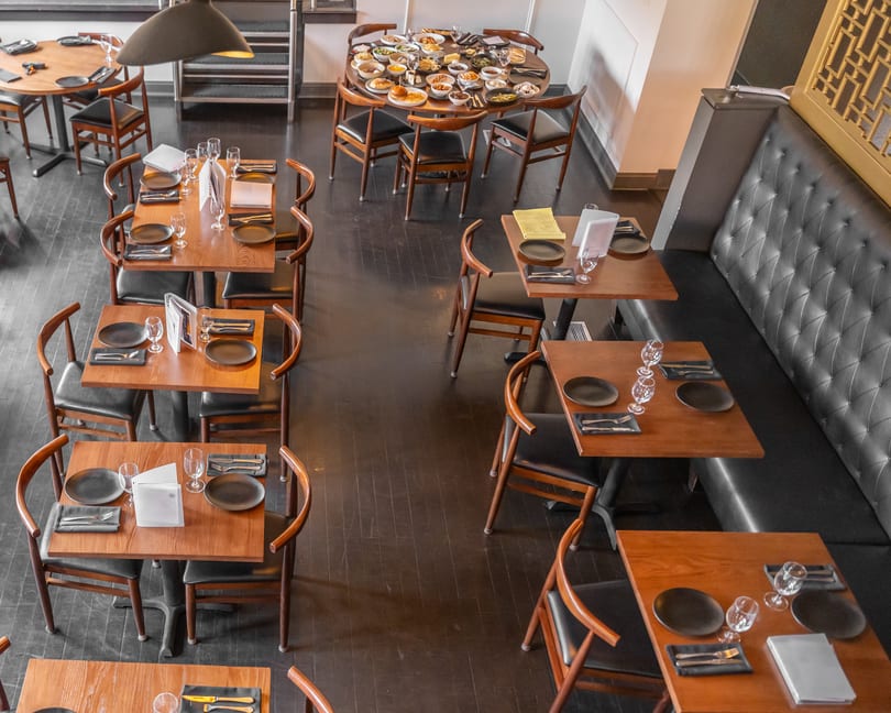 Elegant restaurant dining room with wooden tables, black chairs, a tufted booth, and one table with many small dishes.