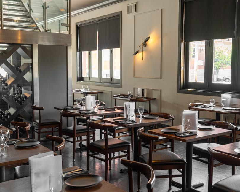 Elegant restaurant interior featuring set tables, dark chairs, large windows, and a decorative wine display.