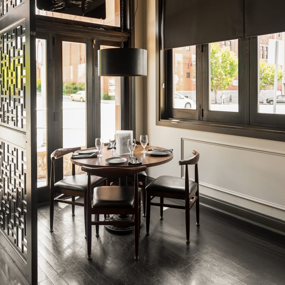 Restaurant dining area with a round wooden table set for four, dark chairs, and large windows.