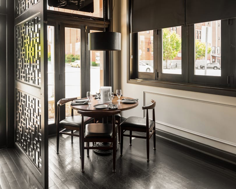 Restaurant dining area with a round wooden table set for four, dark chairs, and large windows.