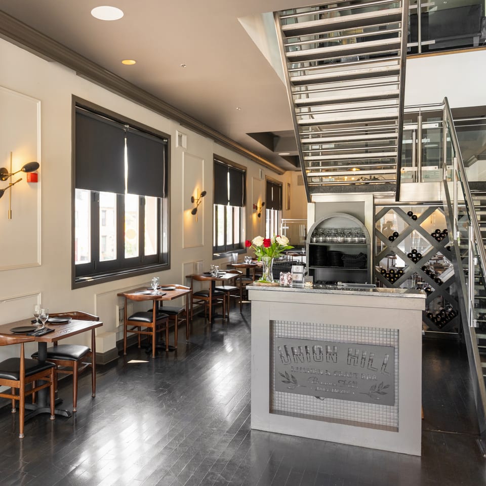 Modern restaurant interior with dining tables, a bar, wine storage, and a sleek metal staircase.