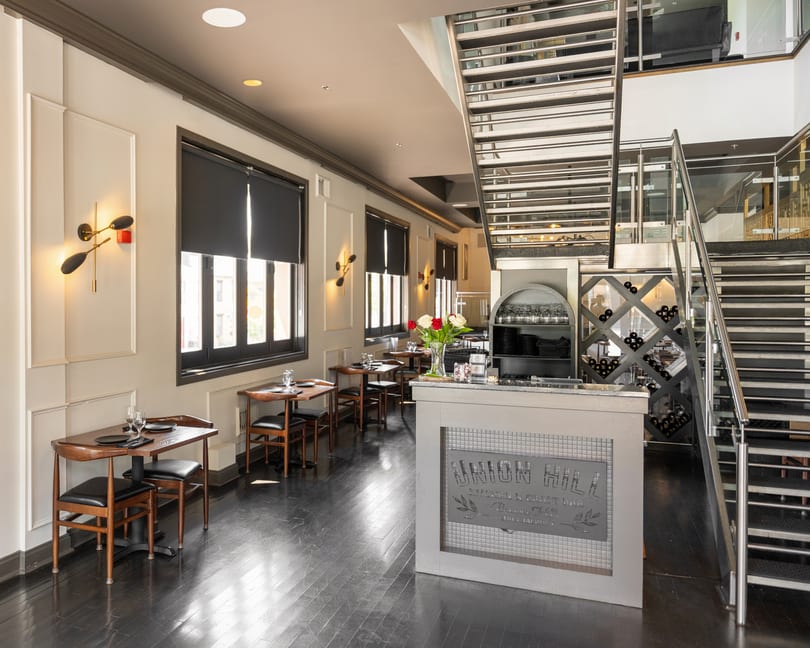 Modern restaurant interior with dining tables, a bar, wine storage, and a sleek metal staircase.