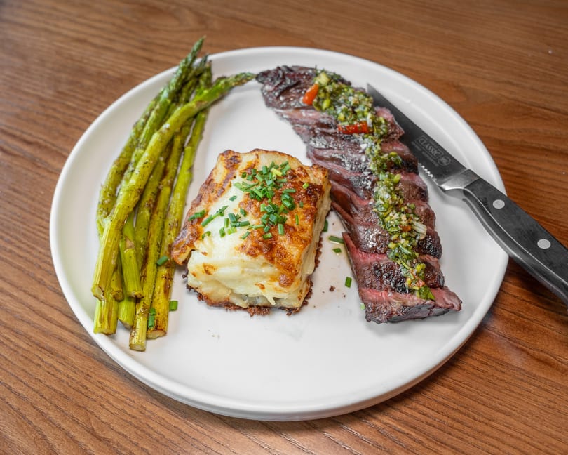 Sliced steak with chimichurri, potato gratin, and asparagus on a white plate with a steak knife.