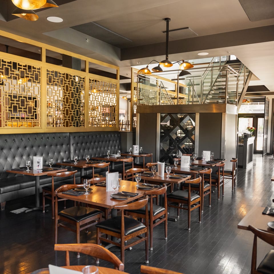 Upscale restaurant dining room with dark wood tables, black booths, golden screens, and a modern staircase.