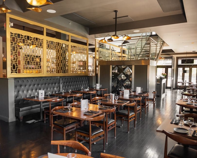 Upscale restaurant dining room with dark wood tables, black booths, golden screens, and a modern staircase.
