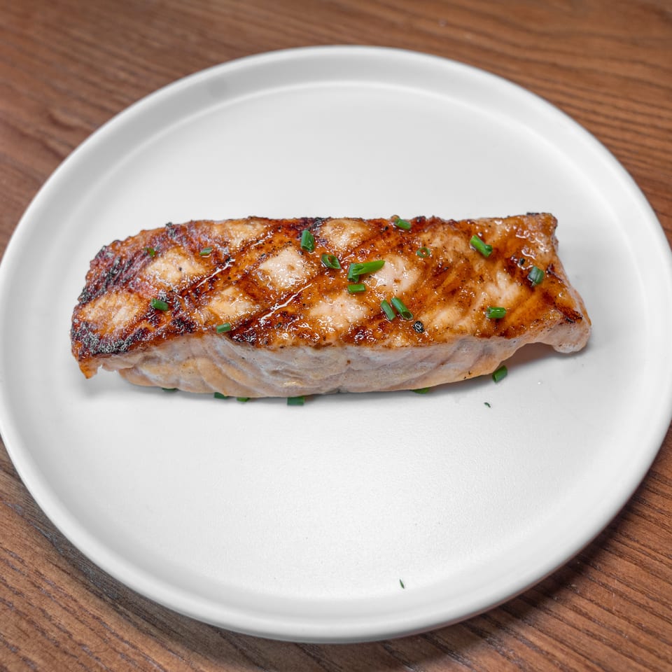 Golden-brown salmon fillet with chives on a white plate, resting on a wooden surface.