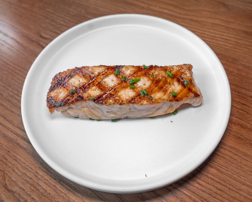 Golden-brown salmon fillet with chives on a white plate, resting on a wooden surface.