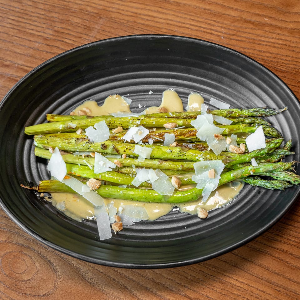 Grilled asparagus spears with creamy sauce, shaved cheese, and chopped nuts on a black plate.