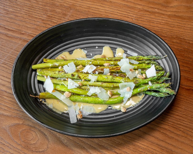 Grilled asparagus spears with creamy sauce, shaved cheese, and chopped nuts on a black plate.