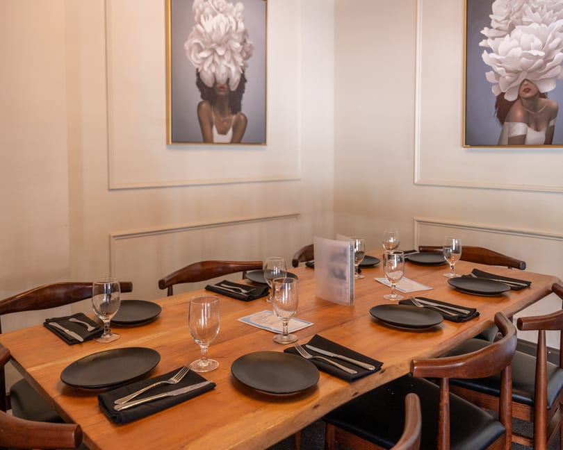 Elegant restaurant dining table set with black plates, glasses, and cutlery, under two framed artworks.