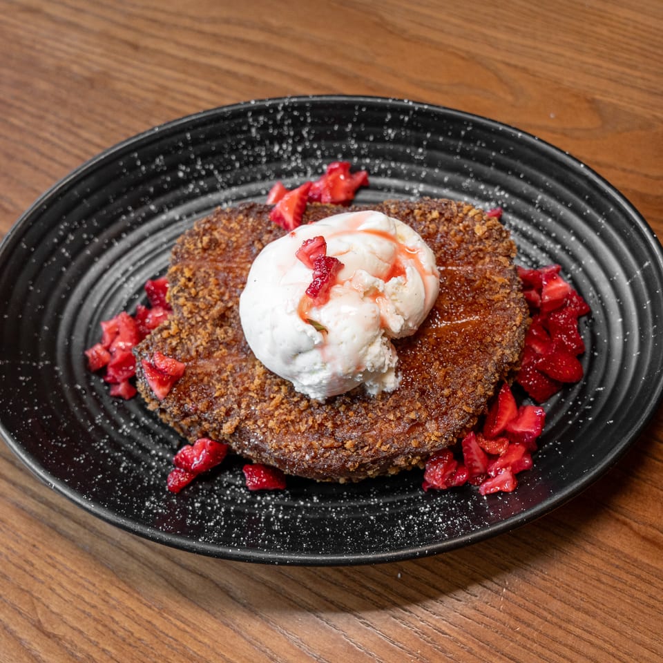 Golden-brown French toast with vanilla ice cream, strawberry topping, and powdered sugar on a black plate.