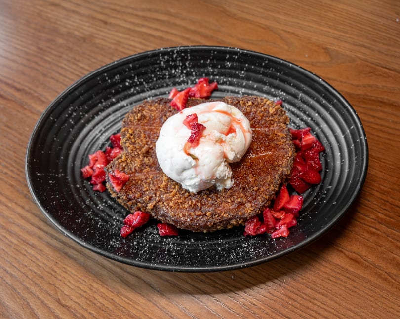 Golden-brown French toast with vanilla ice cream, strawberry topping, and powdered sugar on a black plate.