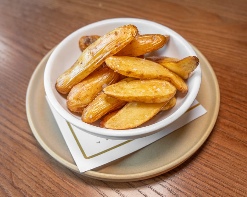 Golden-brown potato wedges sprinkled with salt in a white bowl on a wooden table.